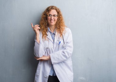 Kameraya bakarak yan el ve parmak ile işaret eden genç redhead doktor kadın yüzüne kocaman bir gülümseme ile bir palto giyiyor gri grunge duvarın üzerinden.