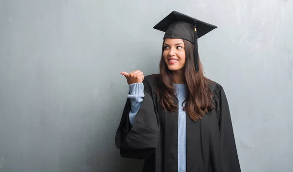 指していると幸せそうな顔を笑顔で側まで親指で示す大学院の制服を着てグランジ灰色の壁を越えて若いブルネットの女性 — ストック写真