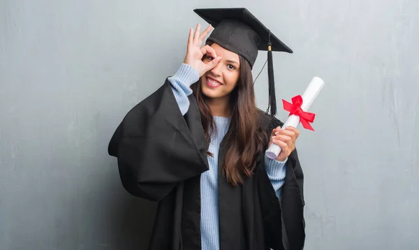 穿灰墙的年轻黑发女子穿着研究生制服持有度与快乐的脸微笑做 标志用手在眼睛看通过手指 — 图库照片
