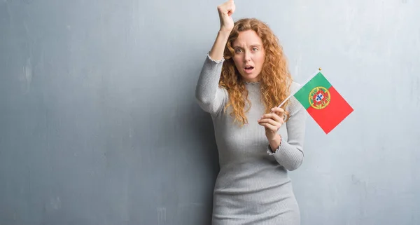 灰色垃圾墙上的年轻红头发的女人拿着葡萄牙国旗懊恼和沮丧的呼喊与愤怒 疯狂和叫喊与举手 愤怒的概念 — 图库照片