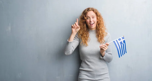 灰色垃圾墙上的年轻红头发的女人持有希腊国旗惊讶的想法或问题指着一个快乐的脸 — 图库照片