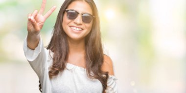 Genç güzel Arap kadın mutlu yüz zafer işareti yaparken kameraya göz kırpıyor ile gülümseyen izole arka plan üzerinde güneş gözlüğü takıyor. İki numaralı.