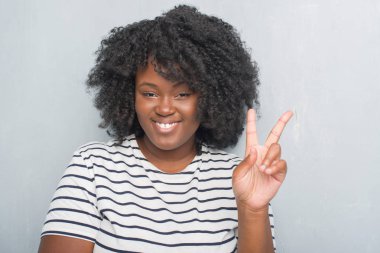 Genç Afro-Amerikan artı boyutu kadın mutlu yüz zafer işareti yaparken kameraya göz kırpıyor ile gülümseyen gri grunge duvarın üzerinden. İki numaralı.