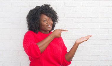 Genç Afro-Amerikan artı boyutu kadın hayrete ve el ile sunulması ve parmak ile işaret ederken kameraya gülümseyen beyaz tuğla duvarın üzerinden.
