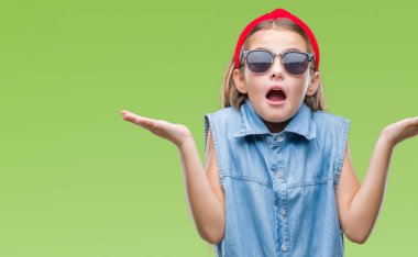 Young beautiful girl wearing sunglasses over isolated background clueless and confused expression with arms and hands raised. Doubt concept. clipart