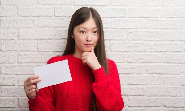 年轻的中国妇女在砖墙上拿着白纸严肃的面孔思考问题 非常困惑的想法 — 图库照片