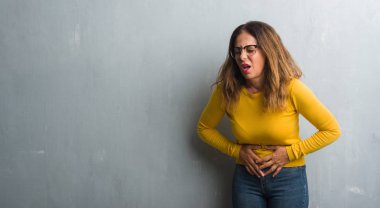 Orta yaş İspanyol kadın el ile mide üzerinde çünkü gözlük gri duvar yüzünden hazımsızlık, ağrılı hastalık hasta hissediyor. Ağrısı kavramı.