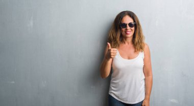 Orta yaş İspanyol kadın gri duvar hareketi el ile mutlu yaşasın yaparken güneş gözlüğü takıyor. Kameraya bakarak başarı gösteren ile ifade onaylama.