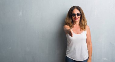 Orta yaş İspanyol kadın gri duvar dostu sunan el sıkışma tebrik ve davet edici olarak gülümseyen güneş gözlüğü takıyor. Başarılı iş.