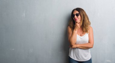 Orta yaş İspanyol kadın gri duvar emin çapraz kollar ve çeneme kaldırdı el ile gülümseme ile kameraya bakarak güneş gözlüğü takıyor. Pozitif düşünce.