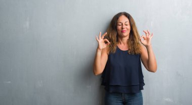 Orta yaş İspanyol gri grunge duvardan duran kadın dinlenmek ve gülümseyen gözlerle parmak ile meditasyon jest yapıyor kapalı. Yoga kavramı.