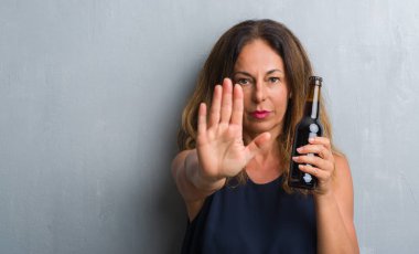 Orta yaş İspanyol kadın açık eliyle dur işareti ile ciddi ve kendine güvenen ifade, savunma hareketi yaparken bira şişesi holding gri grunge duvarın üzerinden ayakta