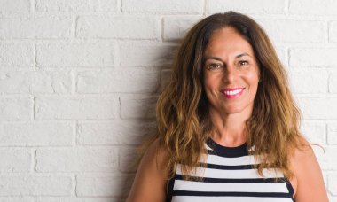 Orta yaş İspanyol kadın ayakta ve diş gösterilen kendine güvenen bir gülümsemeyle gülümseyerek mutlu bir yüz ile beyaz tuğla duvarın üzerinden