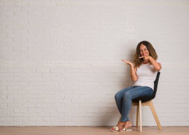 Orta yaş İspanyol kadın beyaz tuğla walll el ve parmak ile işaret çok mutlu üzerinde sandalyede oturuyor