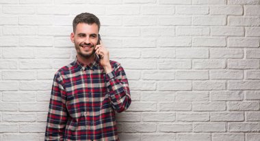 Mutlu yüz ayakta beyaz tuğla duvar üzerinde duran ve diş gösterilen kendine güvenen bir gülümsemeyle gülümseyerek telefonda konuşan genç yetişkin adam