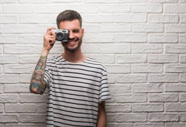 Genç adam ayakta ve diş gösterilen kendine güvenen bir gülümsemeyle gülümseyerek mutlu bir yüz ile beyaz tuğla duvarın üzerinden vintage fotoğraf makinesi ayakta tutan