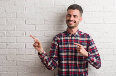 Genç yetişkin adam gülümseyerek ve iki elleri ve parmakları ile tarafına işaret kamera bakıyor beyaz tuğla duvarın üzerinden ayakta.
