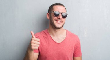Ok işareti, başparmak ile parmak, mükemmel işareti yapıyor kocaman bir gülümseme ile mutlu güneş gözlüğü takıyor gri grunge duvarın üzerinden beyaz delikanlı