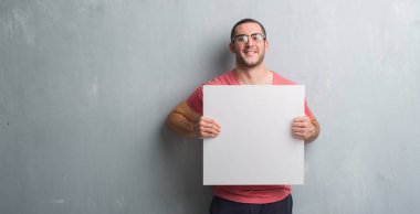 Genç beyaz adam ayakta ve diş gösterilen kendine güvenen bir gülümsemeyle gülümseyerek mutlu bir yüz ile boş bir afiş tutarak gri grunge duvarın üzerinden