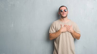 Beyaz delikanlı kapalı gözler ile göğüs ellerinde ve yüzünde minnettar jest ile gülümseyen güneş gözlüğü takıyor gri grunge duvarın üzerinden. Sağlık kavramı.