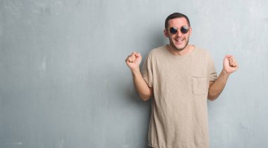 Beyaz delikanlı kutluyor güneş gözlüğü takıyor gri grunge duvarın üzerinden şaşırttı ve başarı için kaldırdı kollarını açarak hayret ve gözleri açık. Kazanan kavramı.