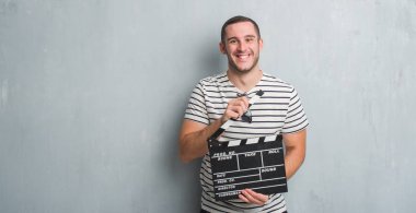 Beyaz delikanlı film fıçı tahtası ile ayakta ve diş gösterilen kendine güvenen bir gülümsemeyle gülümseyerek mutlu bir yüz tutan gri grunge duvarın üzerinden