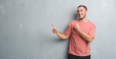 Gülümseyen ve iki elleri ve parmakları ile tarafına işaret kamera bakarak gri grunge duvarın üzerinden beyaz delikanlı.