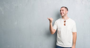 Seyir ve başparmak yanında yukarıyı mutlu yüz ile gülümseyen güneş gözlüğü takıyor gri grunge duvarın üzerinden beyaz delikanlı.