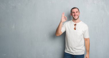 Beyaz delikanlı el ve parmaklar ok işaretiyle yapıyor olumlu gülümseyen güneş gözlüğü takıyor gri grunge duvarın üzerinden. Başarılı ifade.