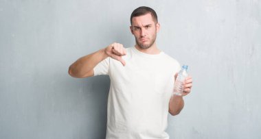 Gri grunge üzerinde beyaz delikanlı duvar holding şişe su kızgın yüzü, eksi işareti aşağı antipati başparmak ile gösterilen, ret kavramı ile