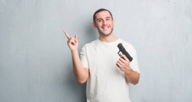 Genç beyaz suç adam elinde silah çok mutlu el ve parmak ile tarafına işaret gri grunge duvarın üzerinden