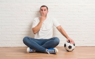 Hata, korku, sessizlik içinde gizli kavramı korkmuş ifade için utanç ile futbol futbol topu kapak ağız eliyle tutarak beyaz tuğla duvar üzerine oturan genç beyaz adam şok