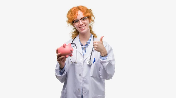 笑顔の サイン 優秀な兆候を親指をやって幸せ貯金箱を保持している赤毛の若い医師女性 — ストック写真