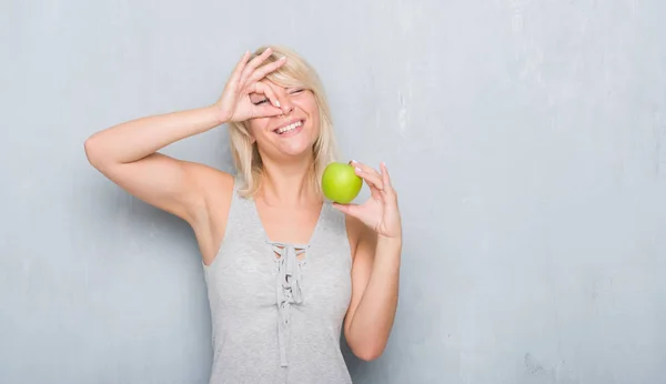 成人白种妇女在垃圾灰色墙壁吃绿色苹果与愉快的面孔微笑做 标志用手在眼睛看通过手指 — 图库照片