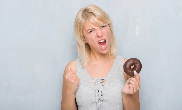 白人成年妇女在灰色的垃圾墙上吃巧克力甜甜圈恼怒和沮丧的呼喊与愤怒 疯狂和叫喊与举手 愤怒的概念 — 图库照片