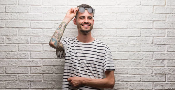 Genç yetişkin adam ayakta beyaz tuğla duvarı görünümlü yan ve dik dik bakma uzağa düşünce gülümseyen güneş gözlüğü takıyor.