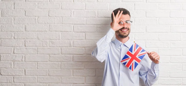指の間から見て目に手で サインをして笑っている幸せそうな顔でイギリスの旗を保持しているレンガの壁を越えて若い成人男性 — ストック写真