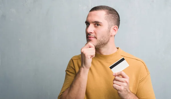 Young Caucasian Man Grey Grunge Wall Holding Credit Card Serious — Stock Photo, Image