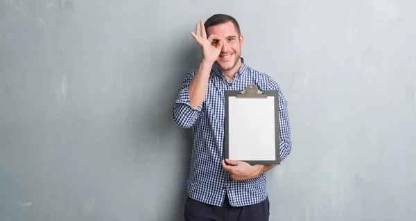 Young Caucasian Man Grey Grunge Wall Holding Clipboard Happy Face — Stock Photo, Image