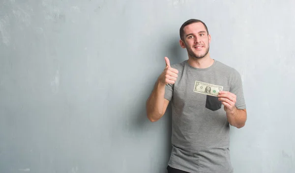 年轻的白种人在灰色垃圾墙显示一美元快乐的大微笑做 拇指与手指 优秀的标志 — 图库照片