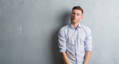 Genç redhead iş adamı gri grunge duvar yapım balık yüzüne dudaklı, çılgın ve komik bir jest. Komik ifade.