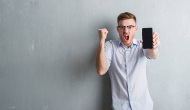 Genç Kızıl saçlı adam gri grunge duvar gösteren boş ekran smartphone rahatsız ve boşa çıkarmak ile öfke, çılgın ve yükseltilmiş eliyle diye bağıran öfke kavramı bağırarak.