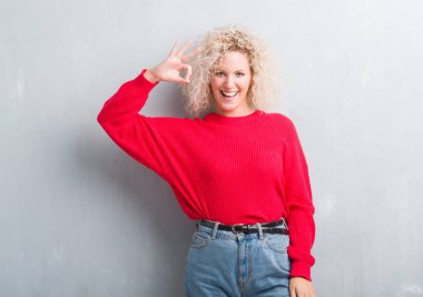 Grunge gülümseyen gri arka plan üzerinde Kıvırcık saçlı el ve parmaklar ok işaretiyle yaparken pozitif sarışın genç kadın. Başarılı ifade.
