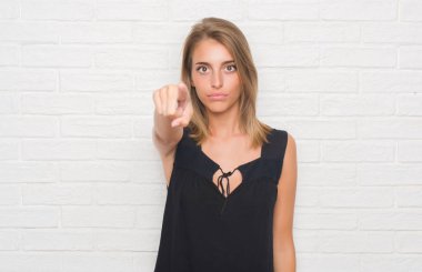 Güzel genç kadın kamera ve size parmak ile işaret beyaz tuğla duvarın üzerinden, el işareti, pozitif ve kendinden emin bir jest ön