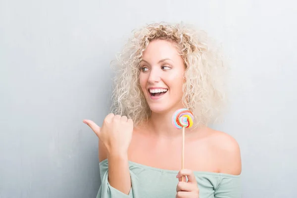 年轻的金发女人在垃圾灰墙上吃糖果指向和显示与拇指向上的一面微笑的笑容 — 图库照片
