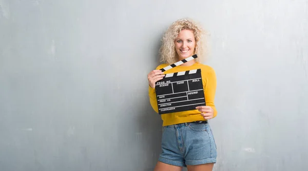 Młoda Blondynka Streszczenie Szary Mur Trzymając Calpboard Filmu Szczęśliwy Twarz — Zdjęcie stockowe