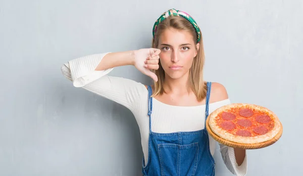 怒った顔 ダウン親指で嫌悪感を示すマイナス記号 拒絶反応の概念でペパロニのピザを食べてグランジ灰色の壁に美しい若い女性 — ストック写真