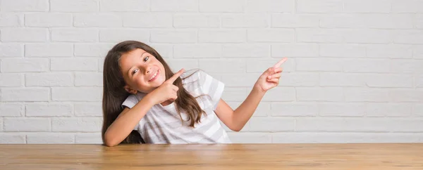 自宅のテーブルの上に座って若いヒスパニックの子供の笑顔と側に つの手と指で指しているカメラ目線 — ストック写真