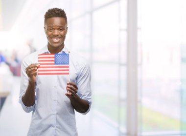 ABD bayrağı ile ayakta ve diş gösterilen kendine güvenen bir gülümsemeyle gülümseyerek mutlu bir yüz tutan genç Afro-Amerikan adam