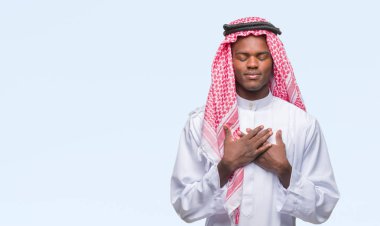 Geleneksel keffiyeh ile gülümseyen izole arka plan üzerinde giyen genç Arapça Afrika adam kapalı gözler ve yüzünde minnettar jest ile göğüs üzerinde eller. Sağlık kavramı.